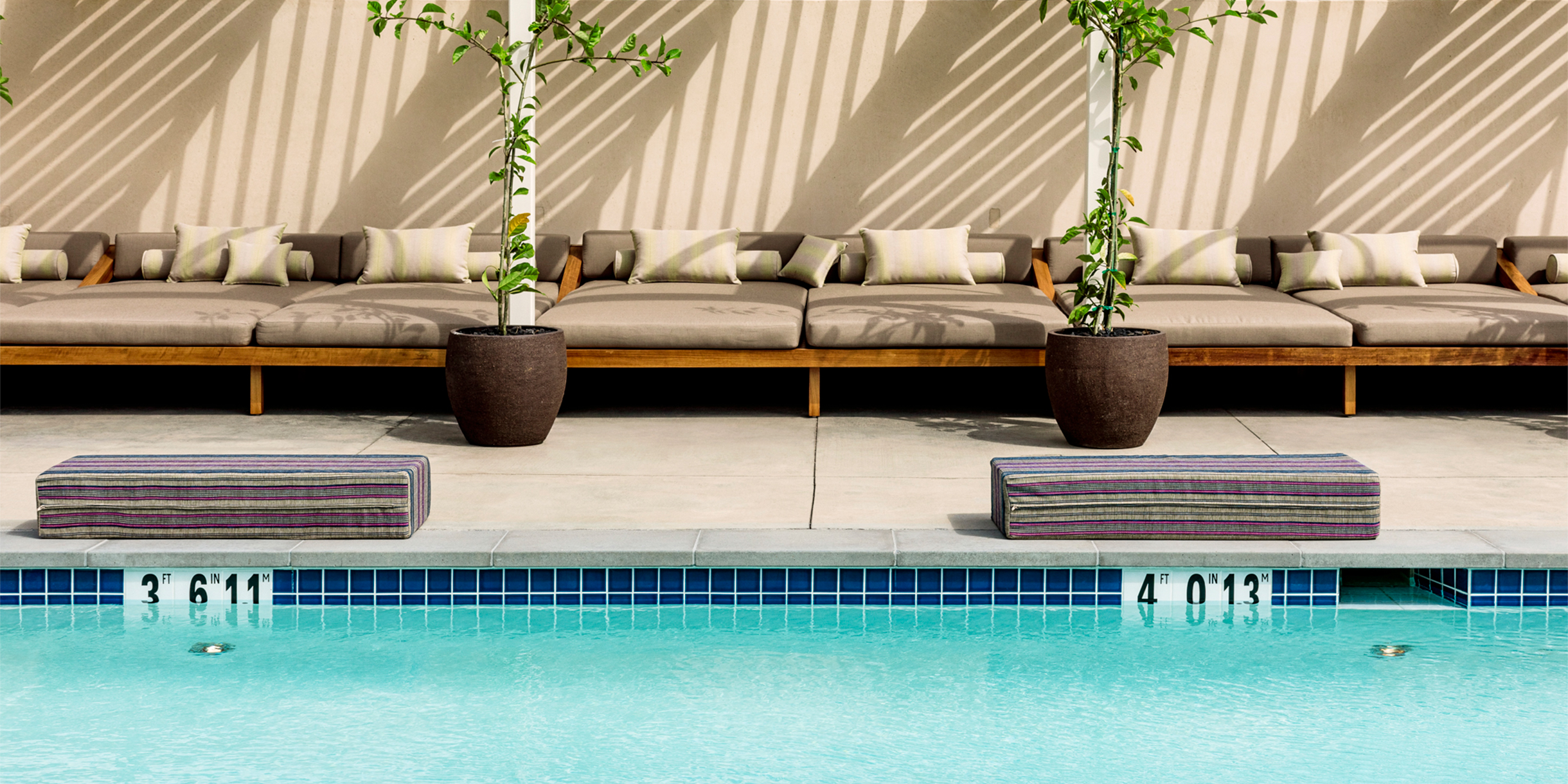 Pool in the foreground, padded cushion next to edge, potted plants and lounge chairs under cabanas