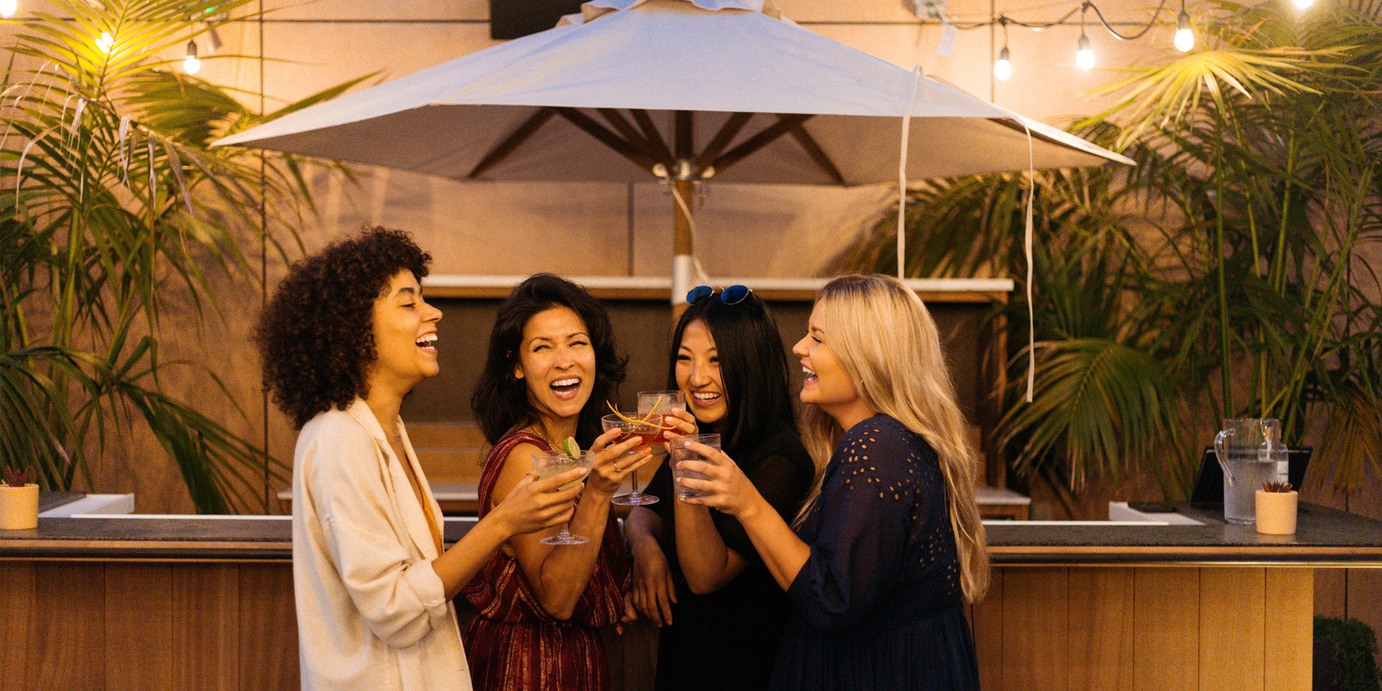 Women laughing, holding cocktails, standing in front of pool bar with umbrella